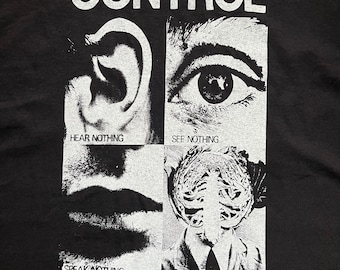 May include: Black and white graphic t-shirt with the word "CONTROL" in large letters at the top. The shirt features three images: an ear, an eye, and a skull. The text "HEAR NOTHING", "SEE NOTHING", and "SPEAK NOTHING" are below the images. The text "CONTROL restrain regulate, authority, means taken to validate experimental results. UNIT a group of things or persons forming a whole.quantity or dimension as a standard of measurement wormwood scrubs and wakofiuld prisons have control units prisoners in these units are subjected to six months tolal sensory deprivation.results from exporinents on prisoners will be adopted as standard measurements with which the authorities will be able to restrain and regulate the group of things or parsons forming the whole of society. THEY MUST BE STOPPED." is at the bottom of the shirt.