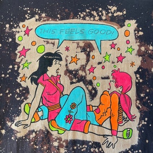 K&ouml;nnte beinhalten: Ein schwarzes T-Shirt mit einem Bleiche-Spritzer-Design, das ein Cartoon-Bild von zwei Frauen mit dem Text "THIS FEELS GOOD!" in einer Sprechblase zeigt.