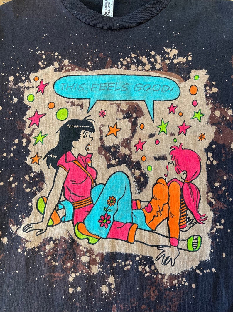 K&ouml;nnte beinhalten: Ein schwarzes T-Shirt mit einem Bleiche-Spritzer-Design, das ein Cartoon-Bild von zwei Frauen mit dem Text "THIS FEELS GOOD!" in einer Sprechblase zeigt.