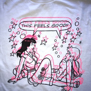 K&ouml;nnte beinhalten: Wei&szlig;es T-Shirt mit einem pinkfarbenen und schwarzen Grafikdruck von zwei Comicfiguren. Die Figuren befinden sich in einer verspielten Pose mit einer Sprechblase, die "This feels good! This feels good!" sagt. Der Hintergrund ist mit schwarzen Sternen und Kreisen gef&uuml;llt.