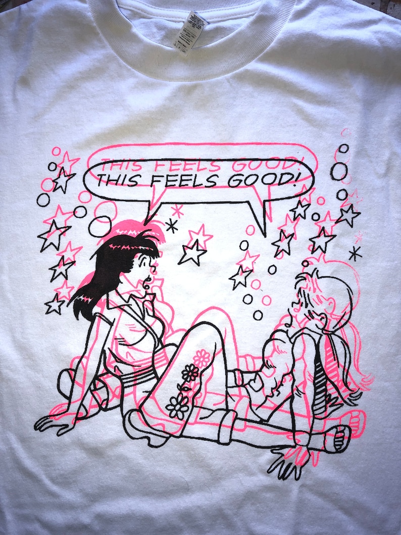 K&ouml;nnte beinhalten: Wei&szlig;es T-Shirt mit einem pinkfarbenen und schwarzen Grafikdruck von zwei Comicfiguren. Die Figuren befinden sich in einer verspielten Pose mit einer Sprechblase, die "This feels good! This feels good!" sagt. Der Hintergrund ist mit schwarzen Sternen und Kreisen gef&uuml;llt.