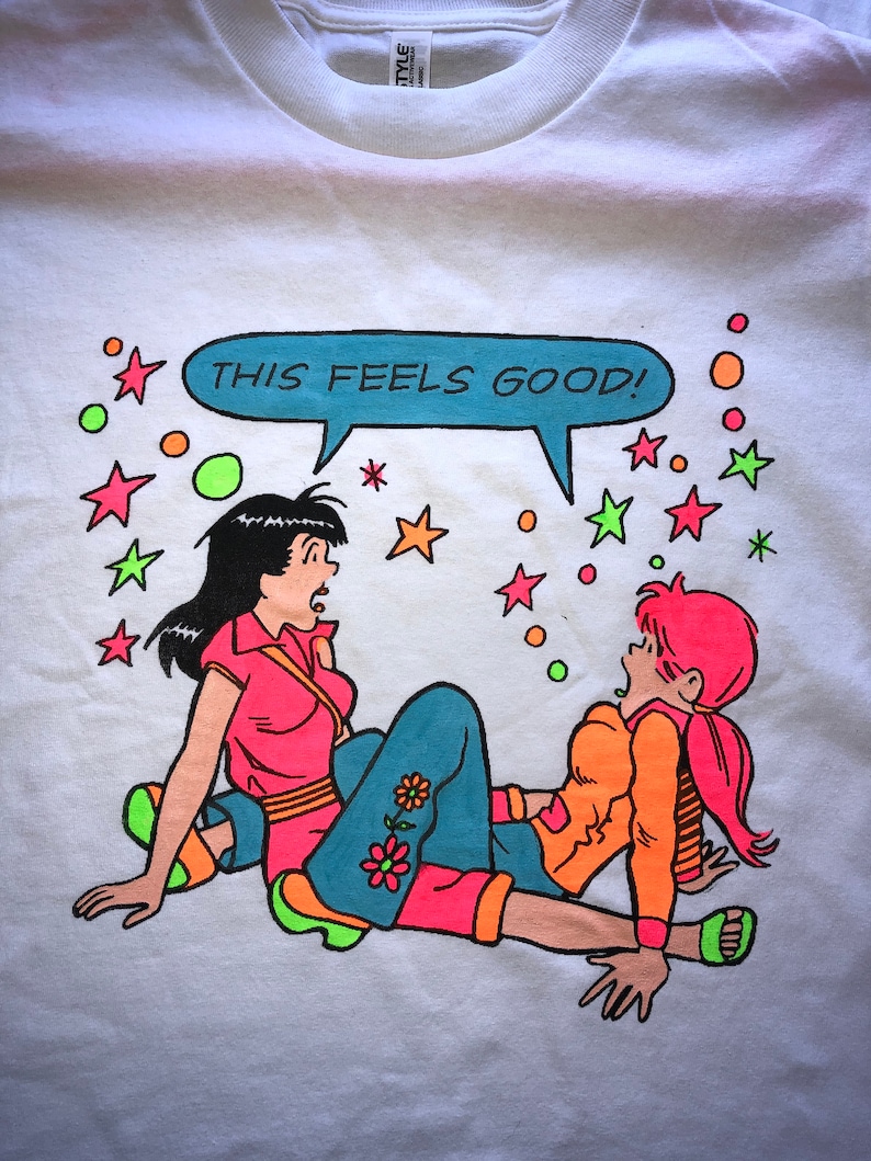 K&ouml;nnte beinhalten: Wei&szlig;es T-Shirt mit einem Cartoon-Motiv von zwei Frauen in bunten Kleidern. Die Frauen sind von Sternen und Kreisen umgeben. Die Sprechblase &uuml;ber ihnen sagt "THIS FEELS GOOD!"