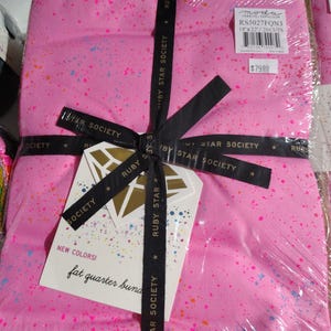 May include: A pink fat quarter bundle with colorful paint splatter design, wrapped in clear plastic and tied with a black ribbon. The ribbon is printed with "RUBY STAR SOCIETY". A tag reads "NEW COLORS! fat quarter bundle".
