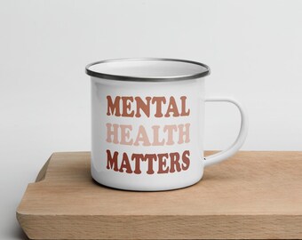 May include: White enamel mug with a silver rim and a handle. The mug features the text "MENTAL HEALTH MATTERS" in a stacked format, using shades of brown. The mug is placed on a wooden surface.