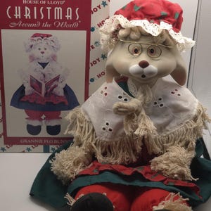 May include: A Granny Flo Bunny doll with a red and white hat, glasses, and a white lace collar. The doll is wearing a red and green dress. A box with the text "CHRISTMAS Around the World" is in the background.