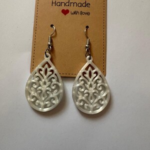 Pode incluir: Um par de brincos brancos em forma de gota com um intrincado design floral vazado. Os brincos penduram de ganchos prateados e são exibidos em uma etiqueta marrom que diz "Handmade with love."