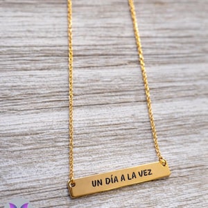 May include: A gold-colored necklace with a rectangular pendant. The pendant has the phrase "UN DÍA A LA VEZ" engraved in black. The necklace hangs on a delicate gold chain against a light wood grain background.