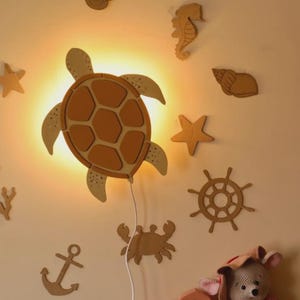 Może przedstawiać: Drewniana lampa ścienna w kształcie żółwia, emitująca ciepłe światło, otoczona morskimi wycinankami z drewna, takimi jak rozgwiazdy, konik morski i koło sterowe. Widoczna jest również pluszowa zabawka myszy z różowym szalikiem i kapeluszem.