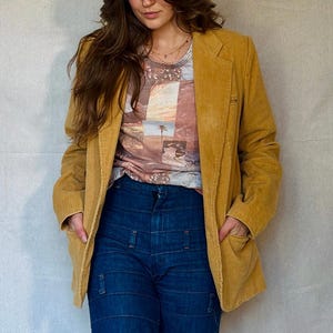May include: A mustard yellow corduroy blazer paired with a graphic tee and dark blue denim jeans. The jeans have a unique patchwork design. The model is wearing a necklace. The outfit is casual and stylish.