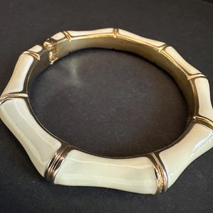 May include: A hinged bracelet with a bamboo design. The bracelet features cream-colored enamel segments with gold-tone metal accents. The bracelet is a bangle style and appears to be closed.