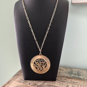 May include: Gold-toned pendant necklace with a circular design. The pendant features an openwork monogram within a larger circular frame. The necklace is displayed on a black mannequin, showcasing the chain and pendant's details. The necklace is a vintage style.