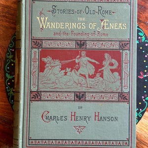 May include: A hardcover book titled Stories of Old Rome: The Wanderings of Aeneas and the Founding of Rome by Charles Henry Hanson. The sage green cover features a central red rectangular panel with a gold illustration of a chariot scene.