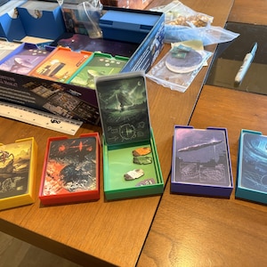 May include: A board game set featuring five colorful plastic card trays in yellow, red, green, purple, and light blue. Each tray holds a deck of illustrated cards with space-themed artwork. A white ruler is visible near the game box.
