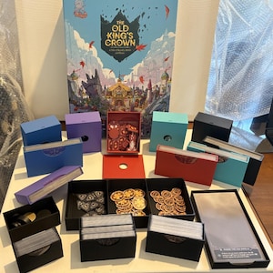 May include: The Old King's Crown board game set includes a large illustrated box, multiple colorful card storage boxes in blue, purple, teal, and black, and several black trays containing round gold-colored tokens and stacks of playing cards.