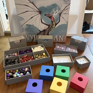 May include: A Wyrmspan board game box featuring a dragon illustration, accompanied by grey plastic card holders, a multi-compartment tray with purple, white, red, and gold tokens, a container of colourful egg tokens, and four square blocks in blue, green, yellow, and red.