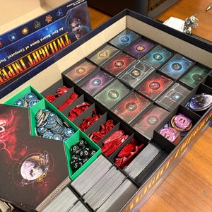 Könnte beinhalten: Geöffnete Brettspielbox mit sortierten Komponenten. Die Box enthält Karten, Spielsteine, Würfel und andere Spielteile. Das Spiel trägt den Titel "Twilight Imperium" und wird als episches Brettspiel über Eroberung, Politik und Handel beschrieben.