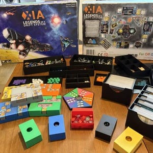 May include: Xia: Legends of a Drift System board game set including two large game boxes, multiple black plastic component organizers, colorful card storage trays, a pentagonal game board piece, and various small game tokens and dice.