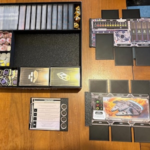 May include: A board game organizer box containing multiple card slots, gold-colored dice, and various circular tokens. Beside the box are two black plastic player dashboards, one featuring a Millennium Falcon card and the other with game stats.