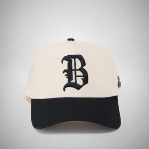 May include: Cream-colored baseball cap with a black brim. The cap features a large, embroidered black letter "B" on the front. A small logo is visible on the side. The hat is a classic style, suitable for casual wear.