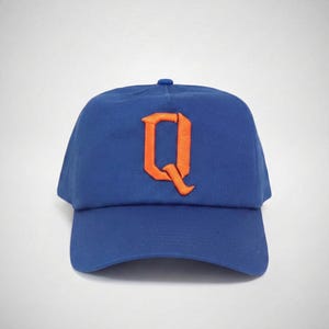 May include: A blue baseball cap with an orange embroidered letter "Q" on the front. The cap has a curved brim and a button on top. The background is plain white.