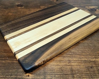 May include: A rectangular wooden cutting board with a dark brown border, a light brown middle section, and a light-colored stripe down the center. The board is made of wood and has a smooth finish. The cutting board is on a wooden surface.