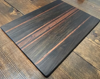 May include: Rectangular wooden cutting board with a dark stained finish. The board features three parallel, lighter-toned wood stripes running across the center. The wood grain is visible, adding texture to the surface. The board is set on a wooden surface.