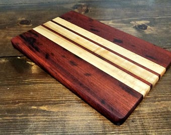 May include: A rectangular wooden cutting board with a rich, dark red-brown hue. It features three parallel stripes of lighter, natural wood running across its surface. The board is likely used for food preparation.