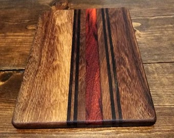 May include: A rectangular wooden cutting board with a mix of light and dark brown wood grains. A central section features a vibrant red stripe, accented by thin black lines. The board's edges are rounded, and the wood has a polished finish.