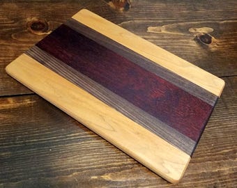 May include: A rectangular wooden cutting board with a striped design. The board features a central section of dark red wood, flanked by bands of gray and light-colored wood. The board is made of multiple wood types.
