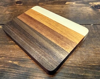 May include: A rectangular wooden cutting board with rounded corners. The board is composed of five parallel strips of wood in varying shades of brown, from dark to light. The wood grain is visible.