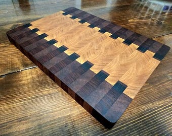 May include: A rectangular wooden cutting board with a unique design. The board features alternating sections of light and dark wood, creating a geometric pattern. The edges are dark, and the board appears to be made of high-quality wood.