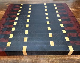 May include: A rectangular wooden cutting board with a dark, almost black center and reddish-brown and black borders. The board features a pattern of small, light-colored rectangular inlays, creating a decorative design. The wood grain is visible.