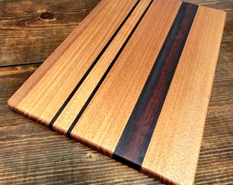 May include: Wooden cutting board with a rectangular shape. The board features a light brown wood grain with two thin black stripes and a wide, dark brown stripe. The board is made of wood.