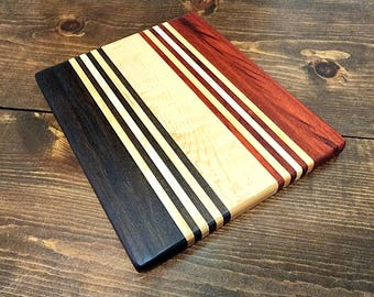 May include: A rectangular wooden cutting board with a striped design. The board features sections of black, light wood, and red-brown wood, separated by thin white stripes. The wood grain is visible, adding texture to the surface.