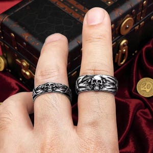 May include: Two silver-toned skull rings. Each ring features a detailed skull design with intricate patterns. The rings are displayed on fingers, showcasing the gothic and edgy style. The background includes a treasure chest and coins.