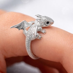 May include: A silver dragon ring with intricate detailing. The dragon has wings, a textured body, and a black eye. The ring is designed to wrap around a finger, showcasing the fantasy-themed jewelry.