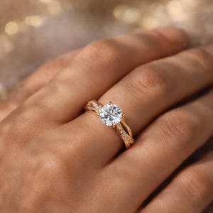 May include: A gold engagement ring with a large, round, clear gemstone. The band has a twisted design with smaller, sparkling stones. The ring is set against a soft, blurred background.