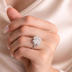 Op de afbeelding: Een zilveren ring met een bloemvormig ontwerp, versierd met meerdere heldere edelstenen. De ring wordt gedragen aan een hand met verzorgde nagels, tegen een zachte, neutrale achtergrond. Het ontwerp van de ring is gedetailleerd en opvallend.
