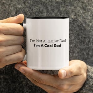 Puede incluir: Taza de cerámica blanca con interior y asa negros. La taza presenta el texto "I'm Not A Regular Dad I'm A Cool Dad" en negro, centrado en la parte delantera. La taza está sujeta por una persona.