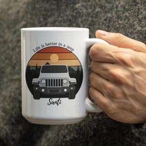 Puede incluir: Taza de cerámica blanca con un gráfico de un Jeep SUV sobre un fondo de puesta de sol, con el texto "Life is better in a jeep" y el nombre "Santi". La taza tiene un asa grande.