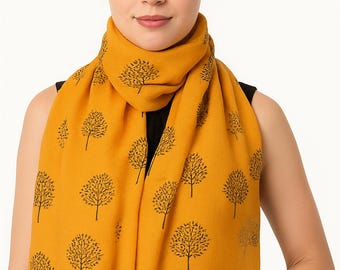 May include: Mustard yellow scarf with a repeating pattern of black tree silhouettes. The scarf is draped around the neck and shoulders. The fabric appears soft and lightweight, suitable for cooler weather. The scarf is a fashion accessory.