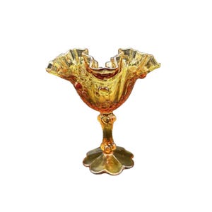 May include: A vintage amber-colored glass bowl with a ruffled rim and pedestal base. The bowl has a textured surface and a scalloped base. The glass is a warm, golden hue, and the design is ornate and decorative.