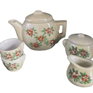 May include: A cream-colored ceramic tea set featuring a teapot, two teacups, a sugar bowl, and a creamer. Each piece is decorated with red floral and green leaf patterns. The teapot has a lid and a handle. The set is designed for serving tea.