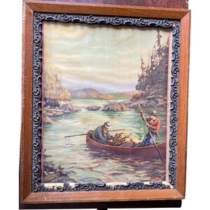 May include: A framed print featuring a landscape painting of two individuals in a canoe on a river. The scene includes a bear on the rocky shore, pine trees, and distant mountains. The frame consists of a smooth light wood outer border and a dark, ornate, embossed inner border with intricate scrollwork patterns.