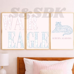 May include: Three framed wall art prints with gold frames. The first two prints feature the text WAR EAGLE in large block letters with a light blue and pink floral pattern. The third print shows a blue tiger illustration and the text AUBURN, ALABAMA.