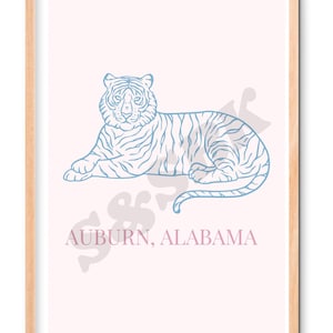 May include: A framed wall art print featuring a light blue line drawing of a tiger lying down on a pale pink background. Below the tiger, the text "AUBURN, ALABAMA" is printed in a light pink font. The print is enclosed in a light wood frame.