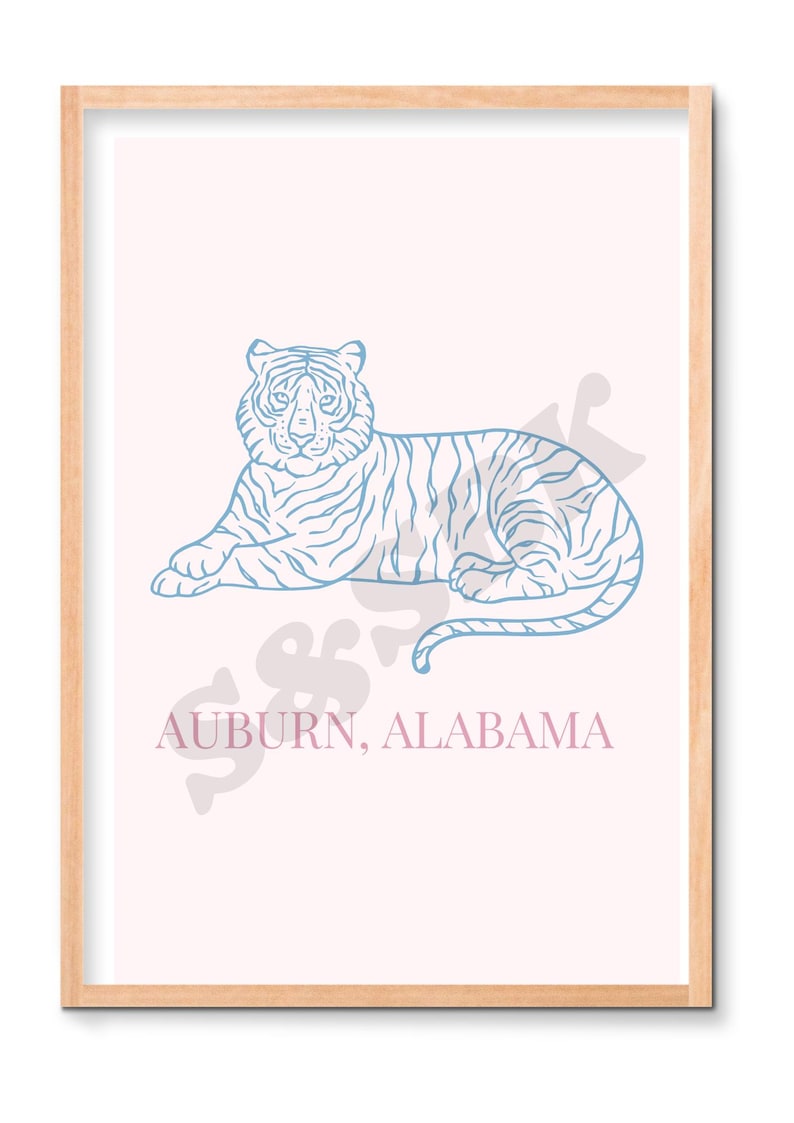 May include: A framed wall art print featuring a light blue line drawing of a tiger lying down on a pale pink background. Below the tiger, the text "AUBURN, ALABAMA" is printed in a light pink font. The print is enclosed in a light wood frame.