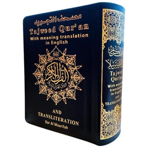 以下が含まれることがあります： Dar Al Maarifah社による「Tajweed Qur'an with meaning translation in English and transliteration」というタイトルの濃紺のハードカバー本。表紙には金色の箔押しでアラビア語のカリグラフィーと、複雑な幾何学模様の星型エンブレムが施されています。