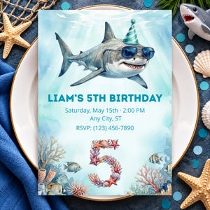 Può includere: Un invito di compleanno rettangolare con uno squalo ad acquerello che indossa un cappellino da festa verde acqua e occhiali da sole blu. Il testo recita LIAM'S 5TH BIRTHDAY, Saturday, May 15th, 2:00 PM, Any City, ST, RSVP: (123) 456-7890, con un grande 5 di conchiglie.