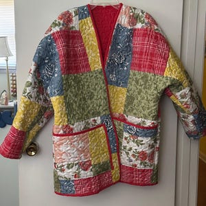 May include: A long-sleeved quilted jacket featuring a patchwork design with various floral and plaid patterns in shades of red, yellow, green, and blue. The garment has a V-neckline, a red trim along the edges, and two front patch pockets.